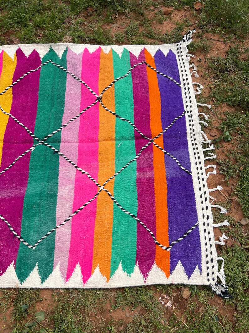 Pode incluir: Um tapete retangular vibrante com listras verticais em amarelo, rosa, laranja, verde, roxo e magenta. O tapete apresenta uma borda branca com um padr&atilde;o em zigue-zague e detalhes em diamante preto e branco.
