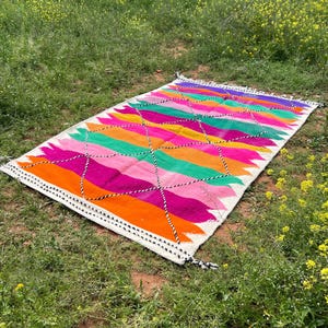 Pode incluir: Um tapete retangular e colorido com um padr&atilde;o geom&eacute;trico. O tapete apresenta listras vibrantes de laranja, rosa, verde, amarelo e roxo. Costuras em preto e branco delineiam o design. O tapete est&aacute; sobre a grama.
