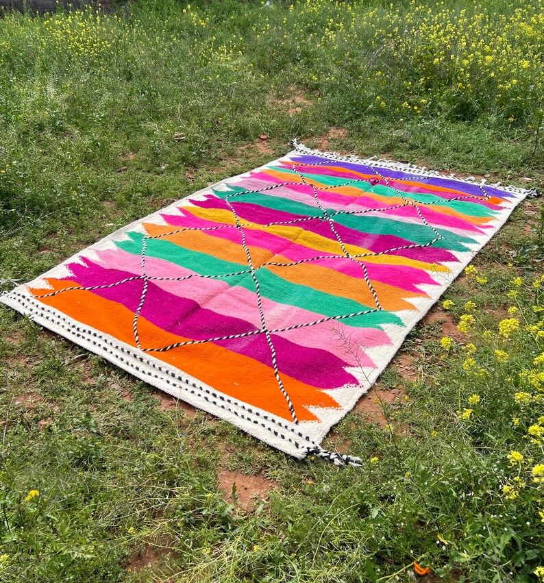 Pode incluir: Um tapete retangular e colorido com um padr&atilde;o geom&eacute;trico. O tapete apresenta listras vibrantes de laranja, rosa, verde, amarelo e roxo. Costuras em preto e branco delineiam o design. O tapete est&aacute; sobre a grama.