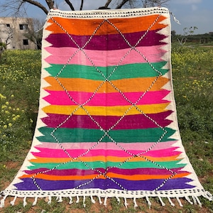 Pode incluir: Um tapete retangular vibrante com um design geom&eacute;trico colorido. O tapete apresenta listras horizontais em tons de laranja, rosa, verde e roxo, intersectadas por um padr&atilde;o de losangos. As bordas s&atilde;o finalizadas com franjas brancas e uma borda branca.