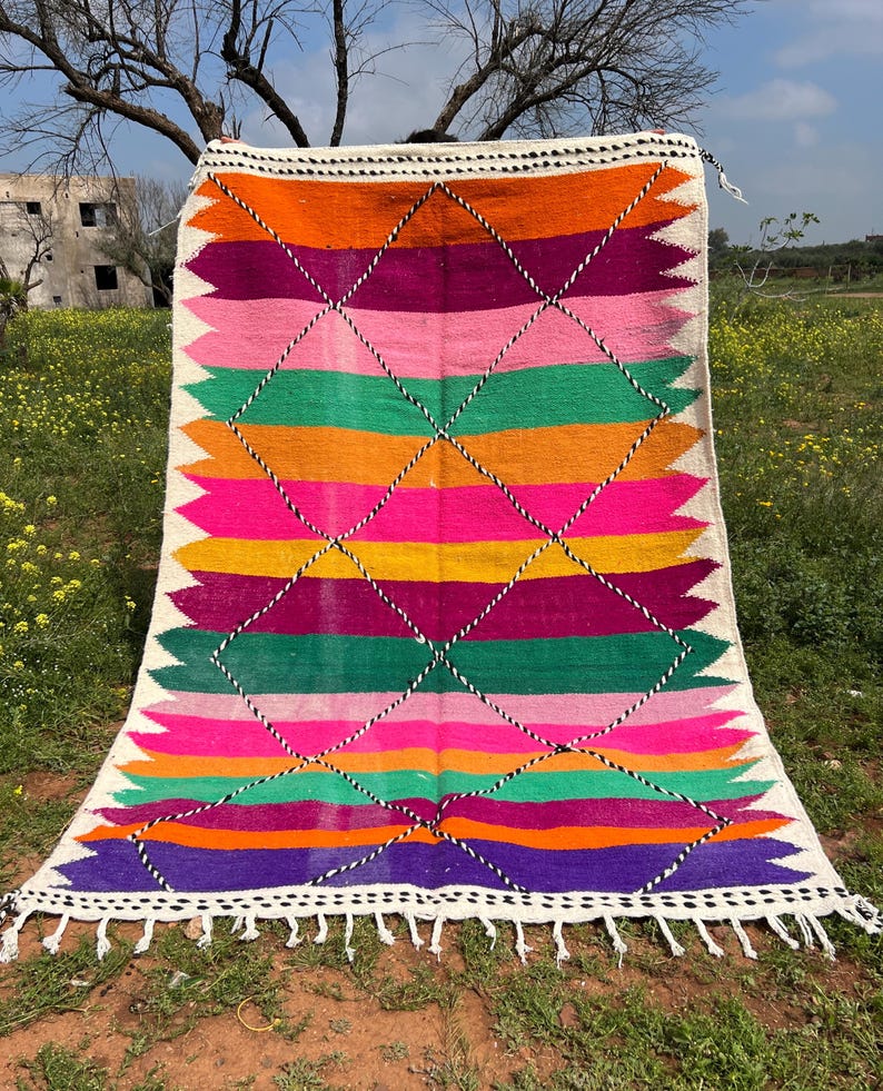 Pode incluir: Um tapete retangular vibrante com um design geom&eacute;trico colorido. O tapete apresenta listras horizontais em tons de laranja, rosa, verde e roxo, intersectadas por um padr&atilde;o de losangos. As bordas s&atilde;o finalizadas com franjas brancas e uma borda branca.