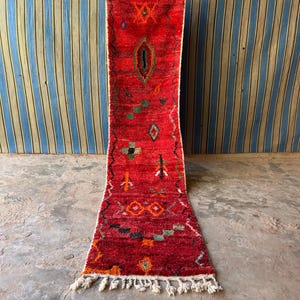 Peut inclure: Un tapis couloir rouge avec des motifs géométriques complexes en orange, vert et noir. Le tapis a une bordure blanche et des franges en bas. Le tapis est présenté verticalement, mettant en valeur son design.