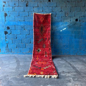 May include: A red, orange and green patterned runner rug with fringe on the ends. The rug is hanging on a blue brick wall.