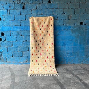 Puede incluir: Una alfombra de color crema con un patrón geométrico de diamantes de colores. La alfombra tiene un borde de flecos y está colgada contra una pared de ladrillo azul.