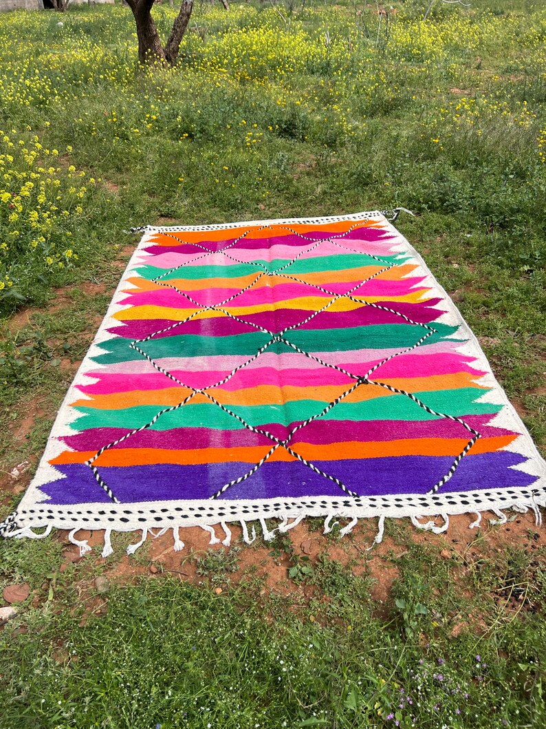 Pode incluir: Um tapete retangular tecido &agrave; m&atilde;o com um design geom&eacute;trico vibrante. O tapete apresenta listras horizontais alternadas em rosa, laranja, verde e roxo, acentuadas por padr&otilde;es de losangos. Possui uma borda branca com borlas. O tapete est&aacute; na grama.