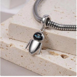 Puede incluir: Un charm de plata con un dise&ntilde;o de robot con cabeza negra y ojos azules, colgando de una pulsera de plata. El charm de robot tiene un cuerpo redondeado y dos brazos peque&ntilde;os. La pulsera tiene un dise&ntilde;o de cadena de serpiente.