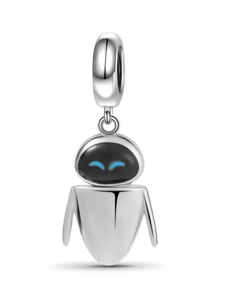 Puede incluir: Un charm de plata con un dise&ntilde;o de robot con cabeza negra y ojos azules. El charm tiene un lazo en la parte superior para sujetarlo a una pulsera o collar. El robot tiene una cabeza redondeada y un cuerpo con lados angulados.