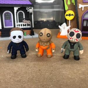 May include: Three small horror-themed figurines standing in a row. The left figure wears a white mask and dark blue jumpsuit. The center figure is a tan burlap sack character in an orange jumpsuit. The right figure wears a hockey mask.