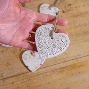 Peut inclure: Ornements blancs en forme de cœur avec des motifs d'oiseaux et de fleurs en relief, attachés avec de la ficelle. L'un est tenu dans une main, d'autres sont visibles. Sur une surface en bois.