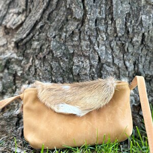 May include: A small, tan leather crossbody bag with a flap made of brown and white fur. The bag is sitting on a bed of green grass.
