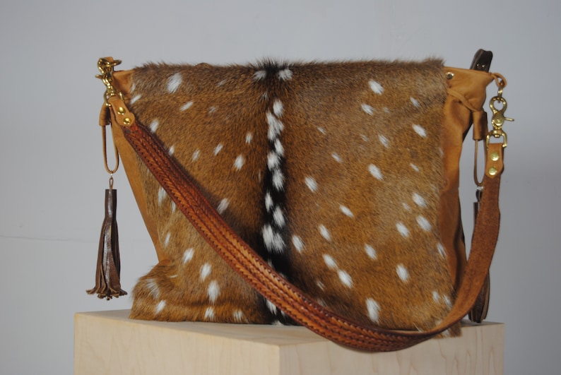 K&ouml;nnte beinhalten: Eine braun-wei&szlig; gefleckte Lederhandtasche mit einem geflochtenen Lederriemen und goldfarbener Hardware. Die Tasche hat eine braune Lederquaste und ein weiches, strukturiertes Aussehen. Die Tasche wird auf einer hellen Holzoberfl&auml;che pr&auml;sentiert.
