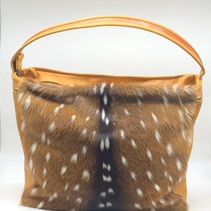 K&ouml;nnte beinhalten: Eine hellbraune Ledertasche mit gefleckter Fellfront. Die Tasche hat einen gebogenen Griff und einen Rei&szlig;verschluss oben. Das Fell ist braun mit wei&szlig;en Flecken, und das Leder ist einfarbig hellbraun.
