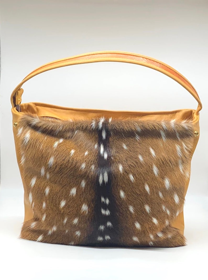 K&ouml;nnte beinhalten: Eine hellbraune Ledertasche mit gefleckter Fellfront. Die Tasche hat einen gebogenen Griff und einen Rei&szlig;verschluss oben. Das Fell ist braun mit wei&szlig;en Flecken, und das Leder ist einfarbig hellbraun.