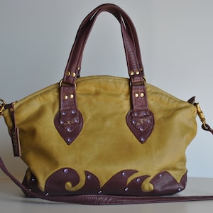 Könnte beinhalten: Eine senfgelbe Ledertasche mit burgunderfarbenem Lederbesatz und einem dekorativen Wellendesign. Die Tasche hat einen Henkel oben und einen langen Schultergurt. Die Tasche hat ein kleines goldenes Etikett mit dem Wort "Marimekko" darauf.