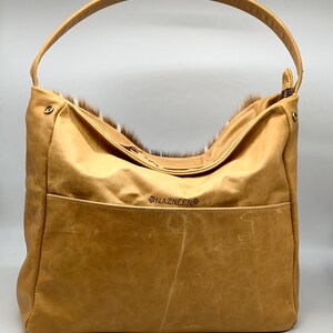 K&ouml;nnte beinhalten: Eine hellbraune Hobo-Tasche aus Leder mit einem einzelnen Henkel. Die Tasche hat eine Vordertasche und eine mit Fell gef&uuml;tterte Oberseite. Der Markenname "NAZNEEN" ist auf der Vorderseite gestickt. Die Tasche ist aus Leder und Fell.