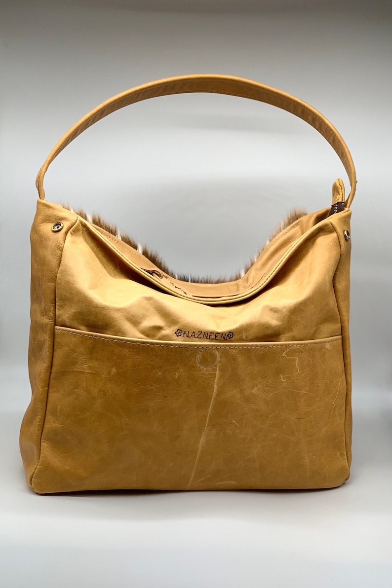 K&ouml;nnte beinhalten: Eine hellbraune Hobo-Tasche aus Leder mit einem einzelnen Henkel. Die Tasche hat eine Vordertasche und eine mit Fell gef&uuml;tterte Oberseite. Der Markenname "NAZNEEN" ist auf der Vorderseite gestickt. Die Tasche ist aus Leder und Fell.