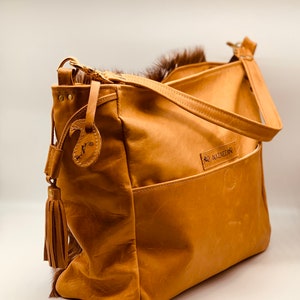 K&ouml;nnte beinhalten: Hellbraune Lederhandtasche mit Schulterriemen, Vordertasche und dekorativem Quastenanh&auml;nger. Die Tasche hat einen Pelzbesatz an der Oberseite und ein kleines Etikett mit dem Buchstaben "N". Der Markenname "NAZREEN" ist auf der Vorderseite sichtbar.
