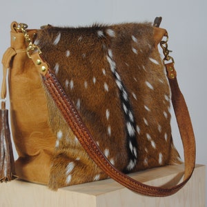 K&ouml;nnte beinhalten: Beigefarbene Leder-Umh&auml;ngetasche mit einem gefleckten Fellbesatz. Die Tasche hat einen geflochtenen Lederriemen, goldfarbene Beschl&auml;ge und eine dekorative Quaste. Die Tasche wird auf einer Holzoberfl&auml;che pr&auml;sentiert.