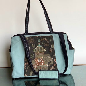 May include: A light blue tote bag with black straps and a large embroidered patch featuring an elephant with a decorative roof on its back. The patch is on a dark brown background with floral designs.