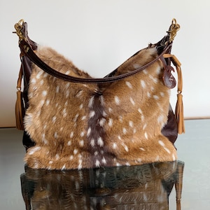 May include: A brown leather handbag with a spotted fawn fur flap. The bag has a long brown leather strap and two tassels.