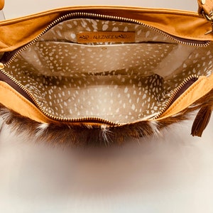 K&ouml;nnte beinhalten: Hellbraune Lederhandtasche mit braunem Fellbesatz, offen gezeigt. Das Innere hat ein geflecktes Musterfutter und ein Lederetikett mit dem Namen "NAZNEEN". Mit Rei&szlig;verschluss und Quastendetail.
