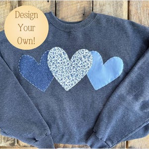 Op de afbeelding: Een donkergrijs sweatshirt met drie blauwe en witte stoffen harten die op de voorkant zijn genaaid. De tekst "Design Your Own!" staat op een cirkel in de linkerbovenhoek van de afbeelding.