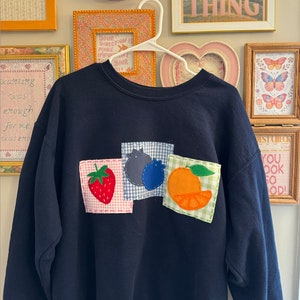 Può includere: Una felpa blu navy con una toppa di fragola, mirtillo e arancia sul davanti. Le toppe sono fatte di tessuto e hanno uno sfondo a scacchi.