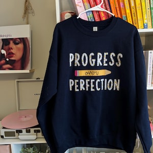 May include: A navy blue sweatshirt with the text "PROGRESS over PERFECTION" in white with a yellow pencil graphic. The text and pencil are decorated with small floral designs.
