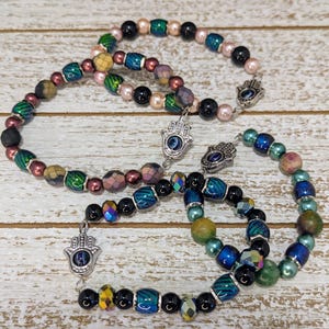 May include: Three beaded bracelets with a variety of colorful beads, including black, pink, green, and iridescent. Each bracelet features a silver Hamsa hand charm with a blue eye design. The bracelets are arranged on a light wood surface.