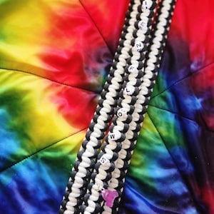 Puede incluir: Dos pulseras de paracord blanco y negro con cuentas blancas que deletrean "FOLLOW" y "ALWAYS". Las pulseras están sobre un fondo de tela de teñido anudado.