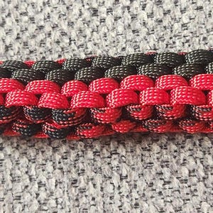 Puede incluir: Un artículo de paracord tejido rojo y negro con un mosquetón rojo y un clip de plástico negro. El paracord está tejido en un patrón repetitivo de rojo y negro. El mosquetón es de color rojo brillante.