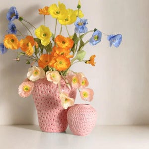 May include: A pink ceramic vase and small matching bud vase, both shaped like strawberries, hold a vibrant arrangement of colorful flowers. The flowers include yellow, orange, blue, and peach blooms. The vases sit on a white surface.