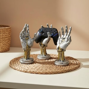 Könnte beinhalten: Drei Roboterhand-Gaming-Controller-Ständer. Die Hände sind silber- und goldfarben, mit komplizierten mechanischen Details. Ein schwarzer Controller ruht auf dem mittleren Ständer. Die Ständer stehen auf einer gewebten Matte.