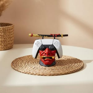 Könnte beinhalten: Ein weiß-schwarzer PlayStation 5-Controller ruht auf einem rot-goldenen Oni-Maskenständer mit einem dekorativen Katana-Schwert. Der Ständer steht auf einer gewebten Matte.
