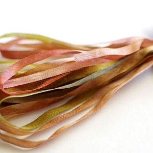 May include: A bundle of thin, multi-colored silk ribbon in shades of pink, brown, and green. The ribbon is arranged in a loose, flowing pile.