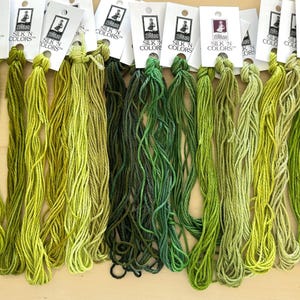 May include: A collection of green silk embroidery threads in various shades, from light yellow-green to deep forest green. Each skein is attached to a white card with the text "SILK N COLORS".