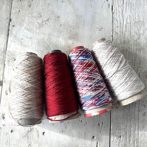 May include: Four spools of thread arranged in a row. The first spool is light beige, the second is solid deep red, the third features a variegated pattern of red, white, and blue, and the fourth spool is a light grey with a subtle textured appearance.