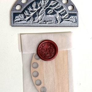May include: A gray wooden half-circle with a carved deer design and holes around the edge. The half-circle is packaged in a white tissue paper envelope with a red wax seal.