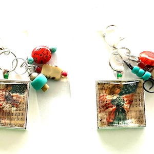 May include: Two square pendants featuring vintage sheet music and American flag imagery, each attached to a silver-tone lobster clasp. The charms are accented with multi-colored beads in shades of turquoise, red, and beige, along with small metal rings.