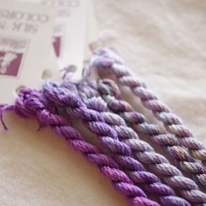 May include: Five skeins of embroidery floss in shades of purple, lavender, and light pink. The floss is arranged in a fan shape and is labeled "Silk N Colors".