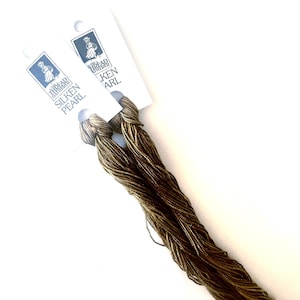 Puede incluir: Dos carretes de hilo Silken Pearl en color oro apagado. El hilo está unido a tarjetas blancas con un logotipo azul y blanco. El hilo está bien enrollado y parece estar hecho de un material fino y sedoso, ideal para bordados u otros proyectos de costura.