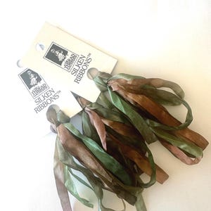 May include: Two bundles of silk ribbons in shades of green, brown, and mauve, with tags that read "Silken Ribbons". The ribbons are tied together, creating a tassel-like appearance. The tags feature a logo of a woman.
