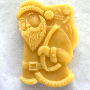 May include: A light yellow, molded Santa Claus figure, carrying a sack over his shoulder. The figure has a long beard, a hat, and a belt with a buckle. The figure is a festive holiday decoration.