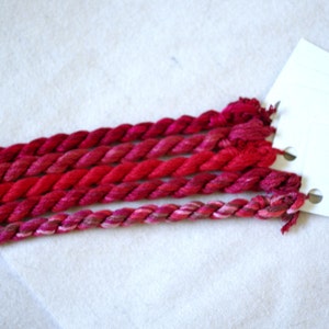 May include: Six skeins of red, pink, and burgundy embroidery floss arranged in a row. The floss is twisted and has a subtle sheen. The skeins are labeled "The Thread Gatherer" and "Silk".