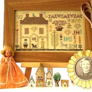 May include: A framed cross-stitch sampler with a house design, a doll in an orange dress, miniature ceramic houses, and a framed portrait. The sampler has the text "A Mother's Heart Leads He Home". A paper heart decoration hangs from the frame.