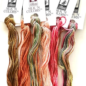 Peut inclure: Gros plan sur six échevettes de fil à broder en soie de différentes couleurs, dont or, orange, rose et vert. Chaque échevette est attachée à une carte blanche avec le texte "SILK 'N COLORS" et "THE THREAD GATHERER".