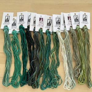 May include: A collection of silk thread skeins in various shades of green, teal, and gray. Each skein is attached to a white card with the text "SILK 'N COLORS". The threads are arranged in a row, showcasing a range of colors.