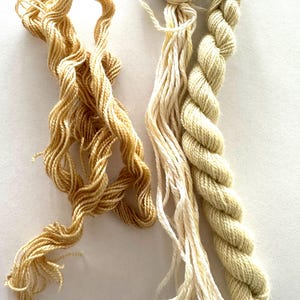 Puede incluir: Primer plano de tres madejas de hilo de bordar en tonos beige y marrón claro. El hilo está bien enrollado y retorcido, con diferentes texturas. Los colores van desde el crema claro hasta un marrón dorado más intenso.