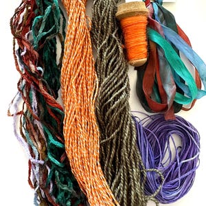 May include: A collection of colorful craft supplies, including various yarns, ribbons, and a spool of orange thread. The yarns are in shades of orange, green, brown, and purple. Ribbons in teal, blue, and burgundy are also present. Ideal for crafting projects.