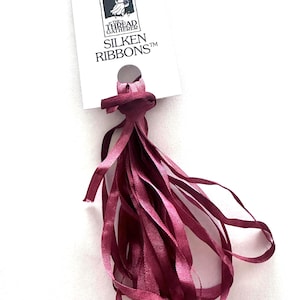May include: A bundle of burgundy silken ribbons hangs from a white tag. The tag features a logo with a woman and the text "THE THREAD GATHERER SILKEN RIBBONS™". The ribbons are long and flowing, with a slight sheen.